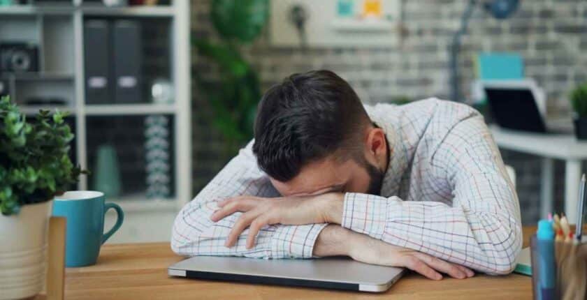 Eine Person sitzt am Schreibtisch und legt den Kopf erschöpft auf die verschränkten Hände.