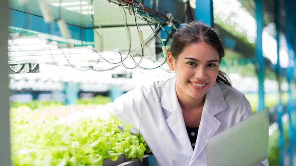 Studentin im Agrarstudium analysiert mit einem Laptop Gemüse auf einer modernen Indoor-Farm.